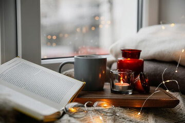 Cup of hot drink, open book and Christmas lights on knitted blanket near window. Cozy season