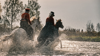 People in old fashion costumes ride on horseback on water and field background. Water drops