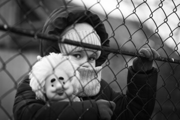 A little refugee girl with a sad look behind a metal fence. Social problem of refugees and...