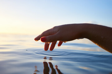 Close-up shot of a female hand touching the surface of calm water at sunset