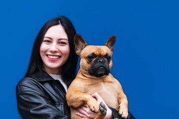 You are my friend! Pleased laughing young asian woman look embraces French Bulldog, expresses true love, cares of favorite pet, isolated over blue background