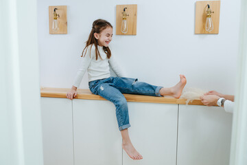 Tween girl sitting on a corridor shelf, her feet being tickled by her mom - obrazy, fototapety, plakaty