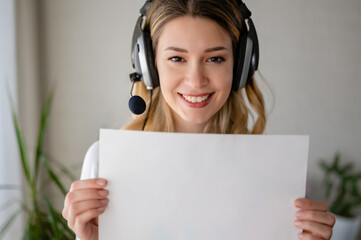 Young beautiful business woman with a headset communicating over the internet, holding a blank paper toward a camera. Mockup