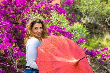 Caucasian woman holding a red umbrella and posing with purple flowers and bushes on the background