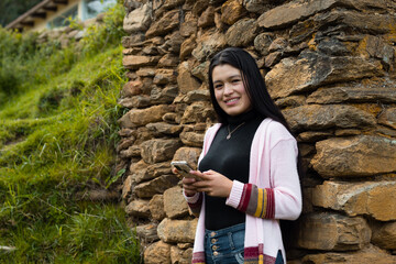 Una atractiva joven con el pelo largo y negro parada cerca de una pared de roca con su móvil en las manos - obrazy, fototapety, plakaty