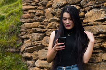 Una hermosa mujer caucásica con cabello largo y negro, felizmente enviando un mensaje a través de su teléfono móvil - obrazy, fototapety, plakaty