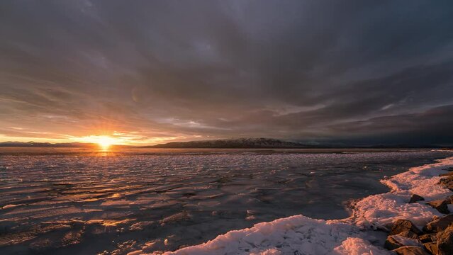 Sun setting over frozen lake as sky lights up with vibrant colors over Utah Lake moving in timelapse.