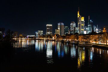 Skyline Frankfurt am Main mit Blick auf den Main von einer Brücke aus. Die Lichter der Büros spiegeln sich auf der Wasseroberfläche. 