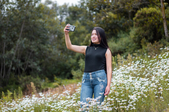 Joven atractiva sonriente  parado tom&aacute;ndose selfie  con su celular en las manos, tel&eacute;fono celular, naturaleza,