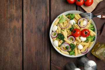 Bowl of delicious pasta with tomatoes, onion and broccoli on wooden table, flat lay. Space for text
