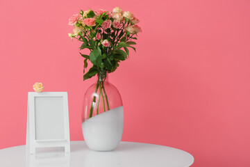 Vase with beautiful roses and frame on table near pink wall