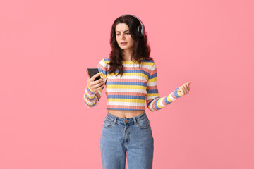 Upset young brunette woman in headphones using mobile phone on pink background