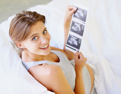 Excited about the little one. A high-angle view of a pregnant woman looking up at the camera while holding a sonogram picture.