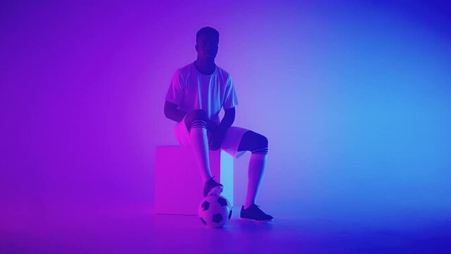 black football forward of halfback is sitting on cube in studio, posing with ball
