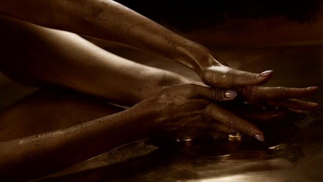 female body in bronze water, woman is smearing gold paint over skin of legs and hands, closeup view