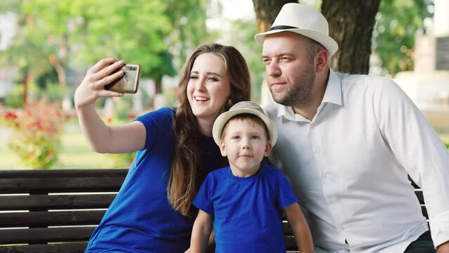 Father toddler mother, parents have fun using smartphone, selfie or video calling together outdoors. Happy family mom dad son, little baby are photographed using modern smartphone in park in summer.