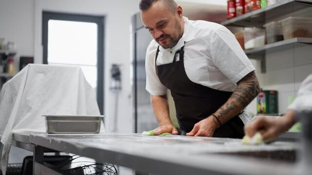Chef and cook cleaning the workspace after doing dishes indoors in restaurant kitchen.
