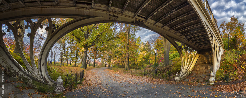 Sticker Panoramic view of a footpath covered by dry leaves pass under a bridge in Central Park in autumn