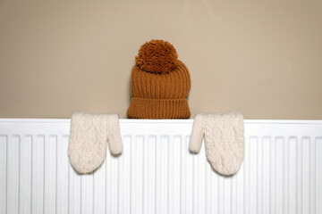 Modern radiator with knitted hat and mittens near beige wall indoors