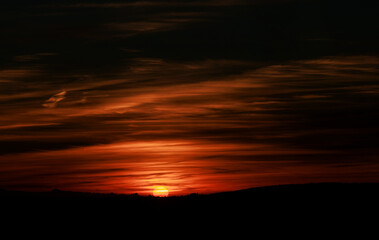 Scenic view of a red sun going down behind the silhouette of the landscape under the cloudy sky