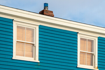 Two double hung windows on a blue exterior wall of a vintage style building. The building has narrow clapboard on the wall. There's a white picket fence in the foreground with a mound of white snow. 