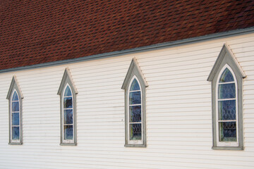 Four clerestory windows in the exterior wall of a white wooden vintage church. The roof of the historic building has red shingles. The windows have grey trim, long edges, and colored stained glass. 