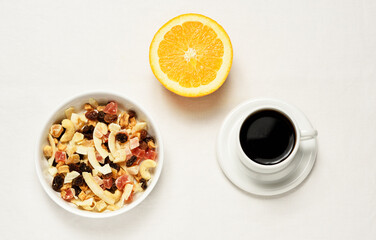 Mixed dried fruit and nuts in a white bowl next to a cut half orange and a cup of black coffee on a white plate