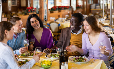 Multiethnic group of positive men and women sitting at table in restaurant, laughing and drinking wine.