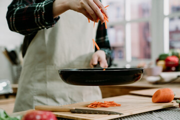 Homemade cooking healthy vegan meal lifestyle concept. unrecognized asian woman in apron putting carrots in hot frying pan with oil. Close up female hands holding fresh vegetables above wooden table