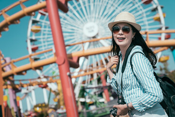 Happy asian chinese young girl backpacker having fun on spring break holiday. Cheerful woman on vacation day tour at ferris wheel. happy backpacker in sunglasses and straw hat carry camera smiling