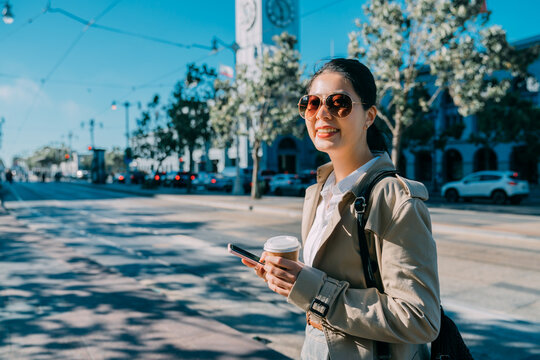 morning coffee take away lifestyle concept. Beautiful young woman hold tea paper cup while walking on street with ferry building in back. cute smiling lady enjoy sunny day using mobile phone outdoor