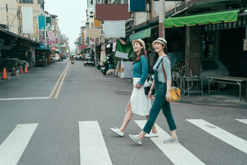 Fototapeta premium side view full length of young female people walking around in town. two smiling girl travelers cross zebra road in taipei taiwan. women traveler relax sightseeing in local traditional market outdoor
