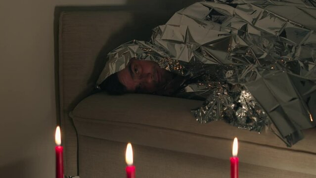 Man covered by a foil blanket lying on couch and looking on lighted candles. Man suffers from electromagnetic hypersensitivity and hides under a metal blanket in dark room with candles. 