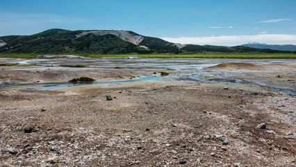 Blue streams from hot springs are visible in the caldera of an extinct volcano. There are sulphurous deposits on the soil. A picturesque mountain range against the azure sky. Kamchatka. Uzon