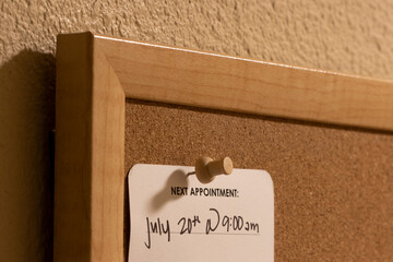 Closeup of a medical appointment reminder card with hand-written date and time on a framed cork board at home.