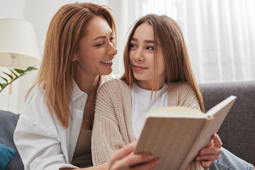 Happy mother and daughter looking at each other while reading book on sofa