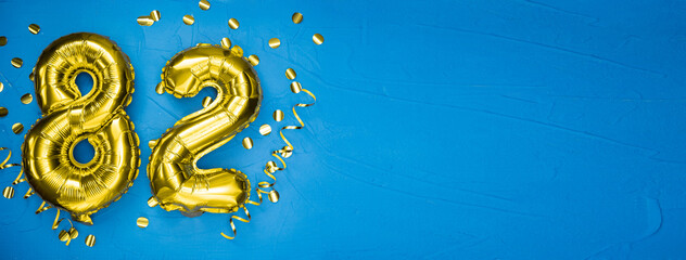 golden yellow foil balloon on blue concrete background number eighty two. Birthday or anniversary card with 82 inscription. Anniversary celebration.