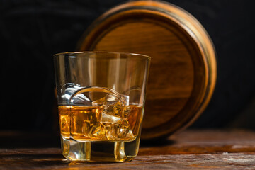 A half-full glass of whiskey with ice cubes stands on dark oak planks against the background of an oak barrel. Close-up. Whiskey from a private distillery.