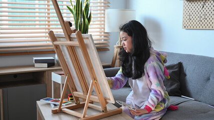 Cute little asian girl is painting picture on easel in living room.