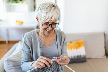 Elderly woman in glasses sit on couch at home smile holding knitting needles and yarn knits clothes...