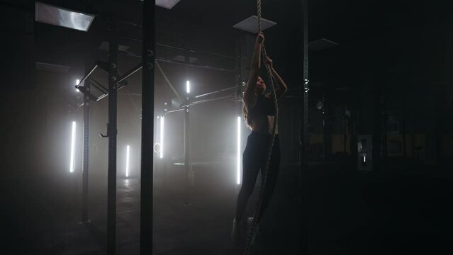 woman is climbing rope in gym, training alone in dark empty fitness hall in evening