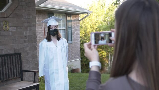 Graduate in 2020 posing for photos in cap and gown with face mask as mom takes photos.