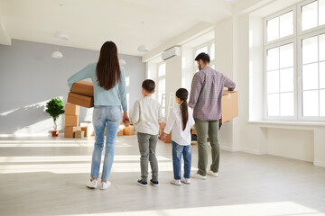 Family buying house or apartment and moving to new home. Back view mom, dad and kids holding cardboard boxes standing in beautiful empty unfurnished spacious living room interior with big white window