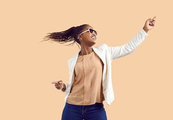 Happy woman vibing to music. Young black girl in suit and sunglasses dancing on solid beige studio...