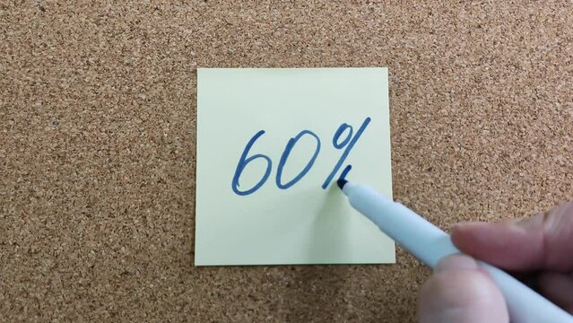 60% with a blue marker on a yellow sticker. Paper sticker on a cork board. Drawing with a marker close-up. Blue felt-tip pen in a woman's hand