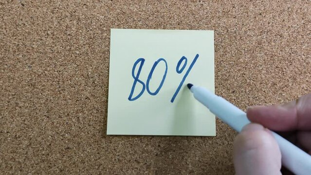 80% with a blue marker on a yellow sticker. Paper sticker on a cork board. A blue felt-tip pen in a woman's hand. Drawing with a marker close-up