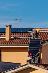 Two workers pass solar panels to each other to be mounted on top of a red tiled roof to install a photovoltaic system