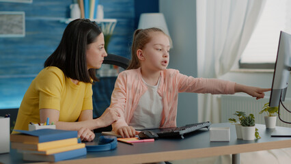 Fototapeta premium Child receiving assistance for online task from mother on computer at home. Parent helping little girl with homework and remote school lessons, pointing at monitor. Schoolgirl and mom