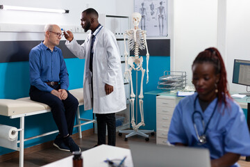 Obraz premium Therapist doctor man measuring temperature using infrared thermometer explaining flu treatment to pensioner man patient during medical consultation in hospital office. Physician scanning forehead