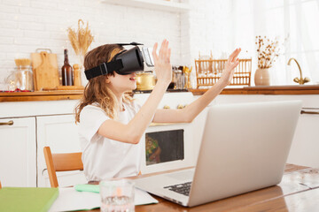 a schoolgirl girl at home in the kitchen with a laptop in vr glasses
